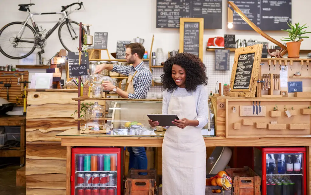 Small business owners in Michigan appreciate the power of Comcast Business and its network to keep mobile devices online with fast, secure and reliable internet. IMAGE: Young African American barista smiling while standing in front of the counter of a trendy cafe using a digital tablet